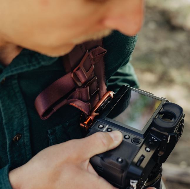 Nahaufnahme eines Peak Design Capture Clips v3 in der edlen Farbvariante Eclipse (Weinrot), montiert am Schultergurt eines Rucksacks. Das Aluminium-Clipsystem hält eine DSLR-/Systemkamera sicher fixiert, während die fotografierende Person das rückseitige