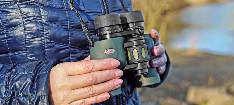 Eine Nahaufnahme einer Person, die ein olivgrünes Kowa YF II Porro-Prismen-Fernglas auf Brusthöhe in den Händen hält. Das Fernglas hängt an einem schmalen, schwarzen Trageriemen um den Hals. Die Hände umschließen den vorderen Bereich des Gehäuses, wobei d