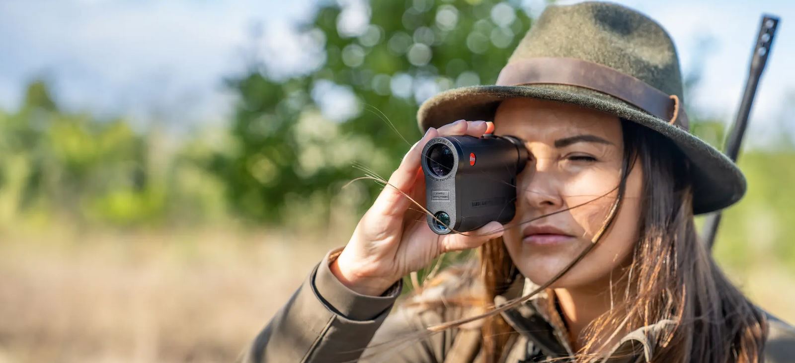 Eine Frau mit einem grünen Jägerhut schaut mit der rechten Hand durch einen Entfernungsmesser der Marke Leica. Sie trägt auf dem Rücken ein Gewehr