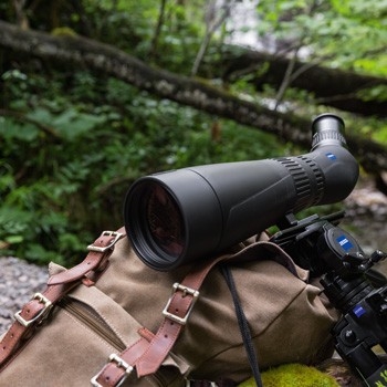 Ein schwarzes Zeiss Victory Harpia 85 Spektiv ist auf einem Stativkopf im Freien montiert und liegt über einem braunen Canvas-Rucksack mit Lederriemen, im Hintergrund ist eine grüne Waldkulisse mit Ästen zu sehen.
