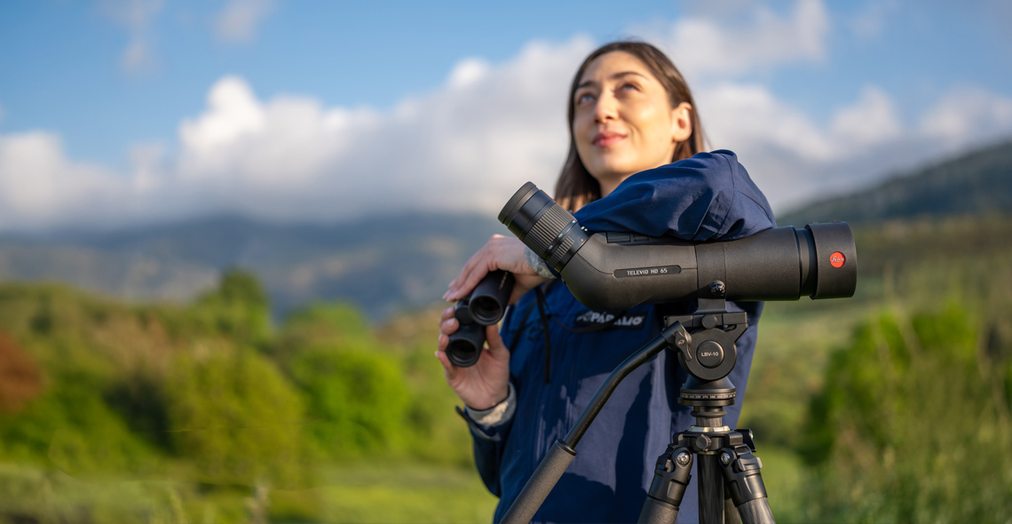 Eine Frau steht in einer grünen Landschaft neben einem Spektiv auf einem Stativ und hält ein Fernglas in der Hand. Unten links steht der Schriftzug „Spektiv-Frühling“.