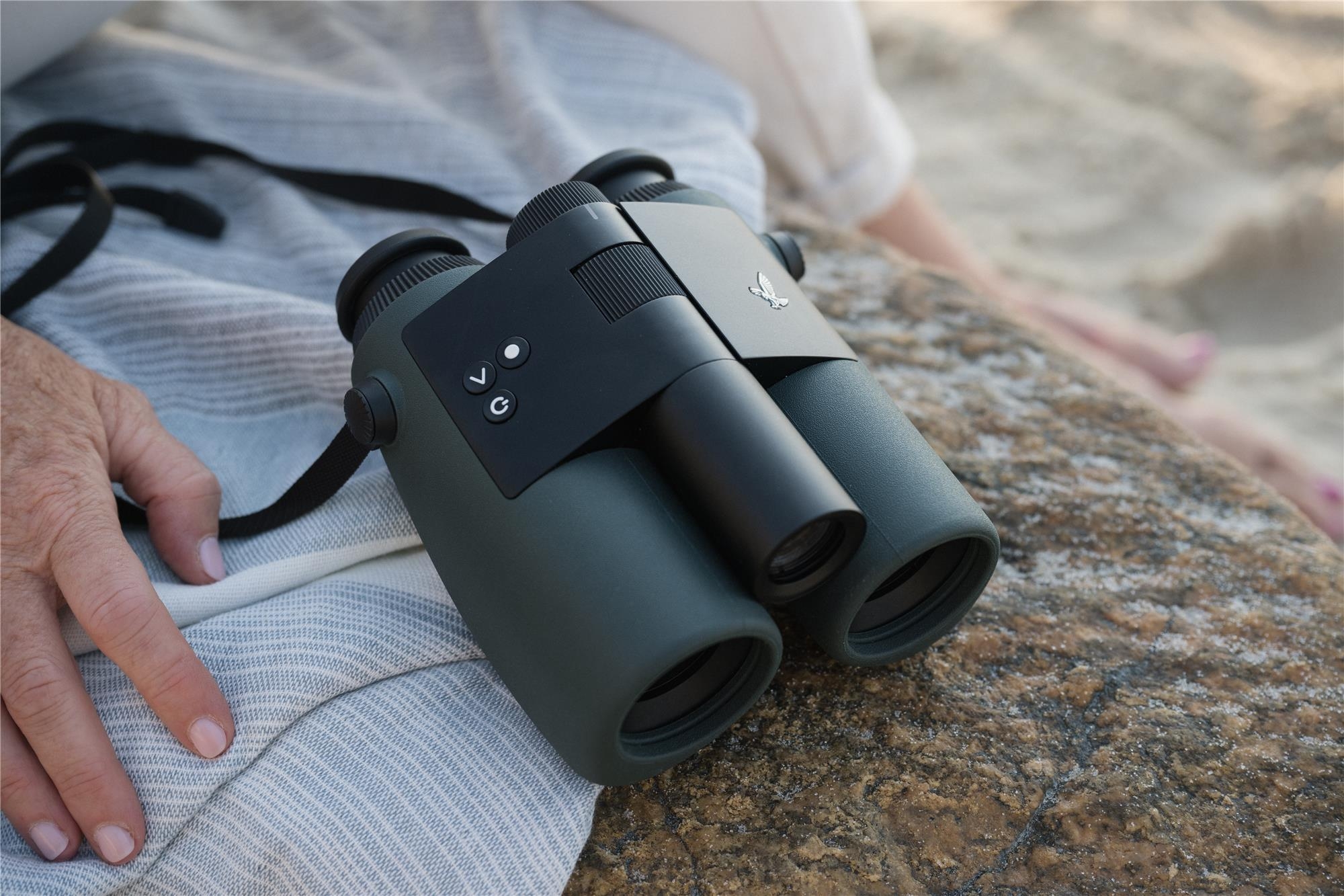 Swarovski AX Visio 10x32 digitales Fernglas in Dunkelgrün mit integrierter Kamera und KI-Objekterkennung, abgelegt auf einem hellen Tuch neben einer Hand im Freien, mit sichtbarem Swarovski-Habicht-Logo auf der Brücke und Funktionstasten am Gehäuse.