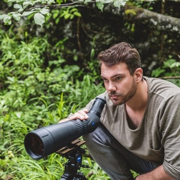 Ein Mann blickt konzentriert durch das ZEISS Victory Harpia 85 Spektiv mit Schrägeinblick, das auf einem Stativ montiert ist, inmitten einer grünen, dicht bewachsenen Waldumgebung.