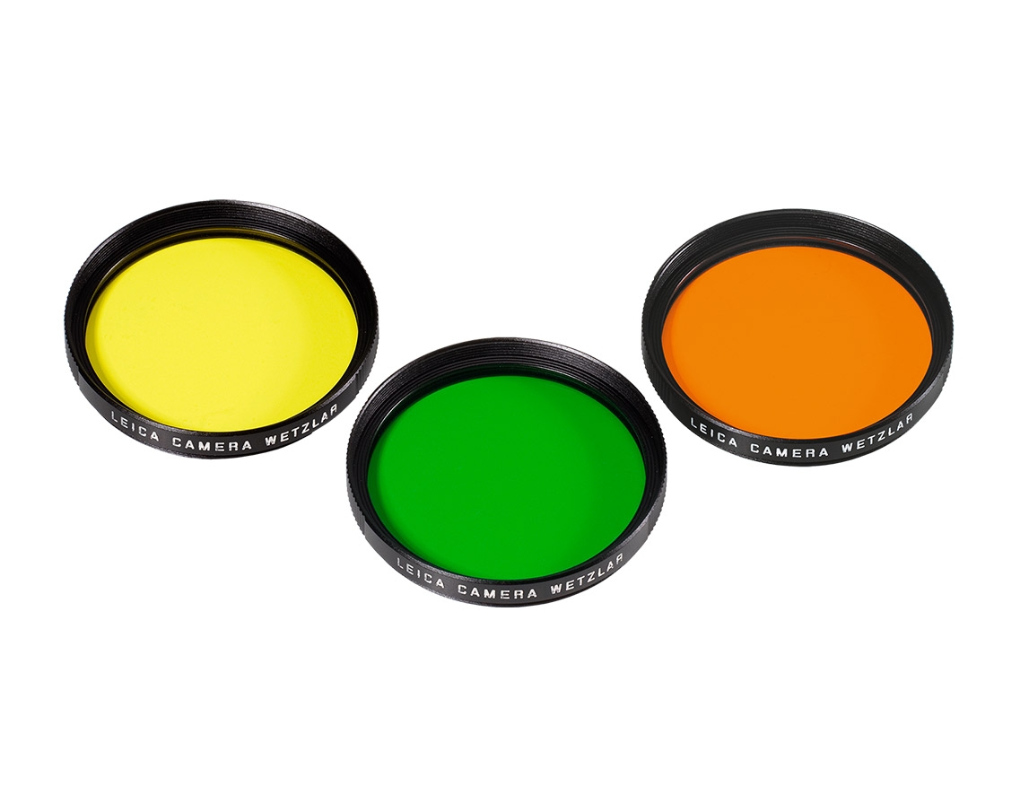 Perspektivische Draufsicht eines dreiteiligen Leica Objektivfilter-Sets mit einem gelben, einem grünen und einem orangefarbenen Kontrastfilter für die Schwarzweißfotografie mit schwarzem Metallrahmen vor weißem Hintergrund.