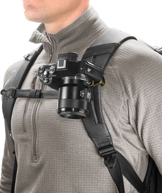 Eine Person im Profil von vorne, die eine schwarze Systemkamera mittels einer Peak Design Capture Clip v3 Kamerahalterung am linken Schultergurt eines Rucksacks befestigt hat. Die Halterung fixiert das Kameragehäuse mit angesetztem Objektiv stabil und gri