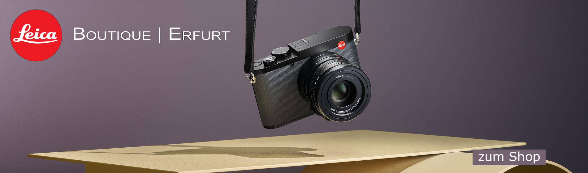 Der Hintergrund ist flieder-farben, links oben ist das Leica Logo und in weißer Schrift steht daneben Boutique Erfurt. Eine Leica Kamera hängt in der Mitte an einem Gurt über einem Birkenfarbenem Tisch. Unten rechts steht ein Button mit weißer Schrift: zu