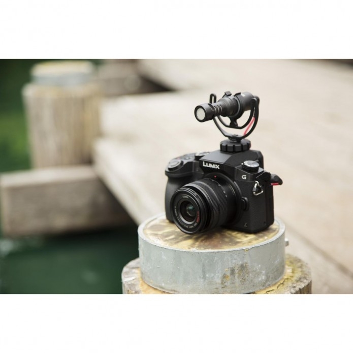 Eine schwarze Panasonic Lumix G7 Systemkamera mit dem montierten Røde VideoMicro Richtmikrofon steht im Freien auf einem runden Holzpfosten an einem Steg im unscharf verlaufenden Hintergrund (Bokeh-Effekt).