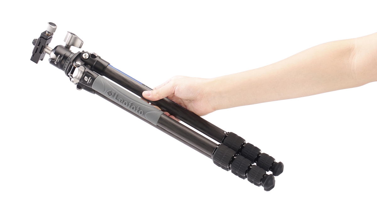 Eine Hand hält das kompakt zusammengeklappte, leichte Leofoto Ranger Carbon-Stativ mit Kugelkopf schräg vor weißem Hintergrund, um das geringe Gewicht und die handliche Größe (Packmaß) für die Reisefotografie zu demonstrieren.