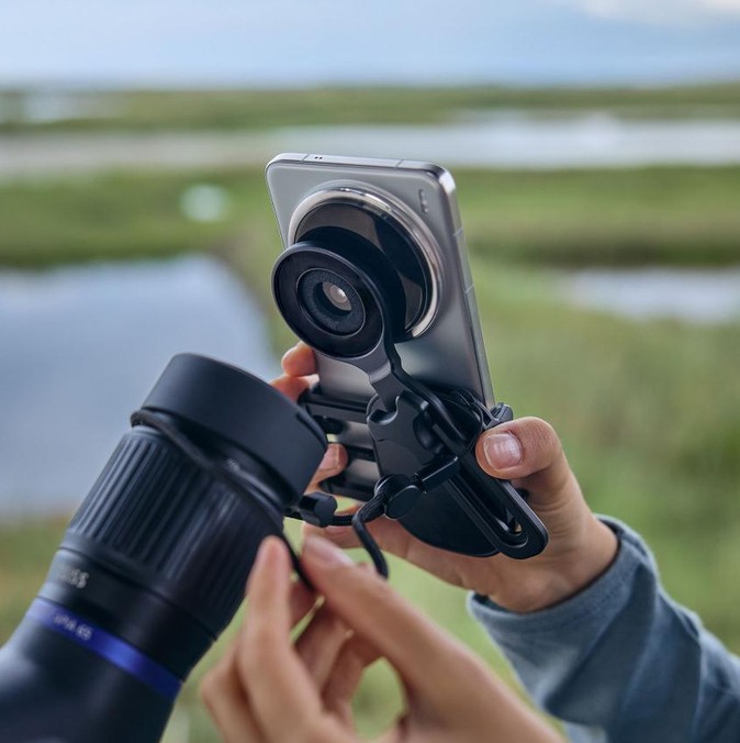Eine Person hält im Freien ein Smartphone in einer Halterung, an der ein runder Adapterring über der Kameralinse angebracht ist. Mit der anderen Hand wird das Smartphone-Konstrukt an das Okular eines ZEISS Spektivs oder Fernglases herangeführt, um ein Fot