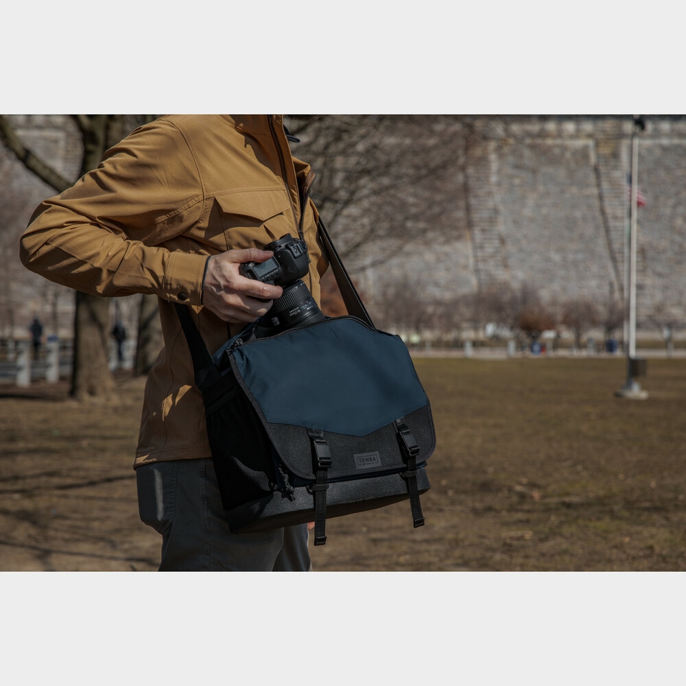 Eine Außenaufnahme einer Person in einer braunen Jacke, die eine spiegellose Systemkamera aus einer blau-schwarzen Tenba DNA Messenger-Kameratasche zieht. Die Tasche hängt an einem Schultergurt über dem Körper und ist mit geöffnetem oberen Reißverschluss