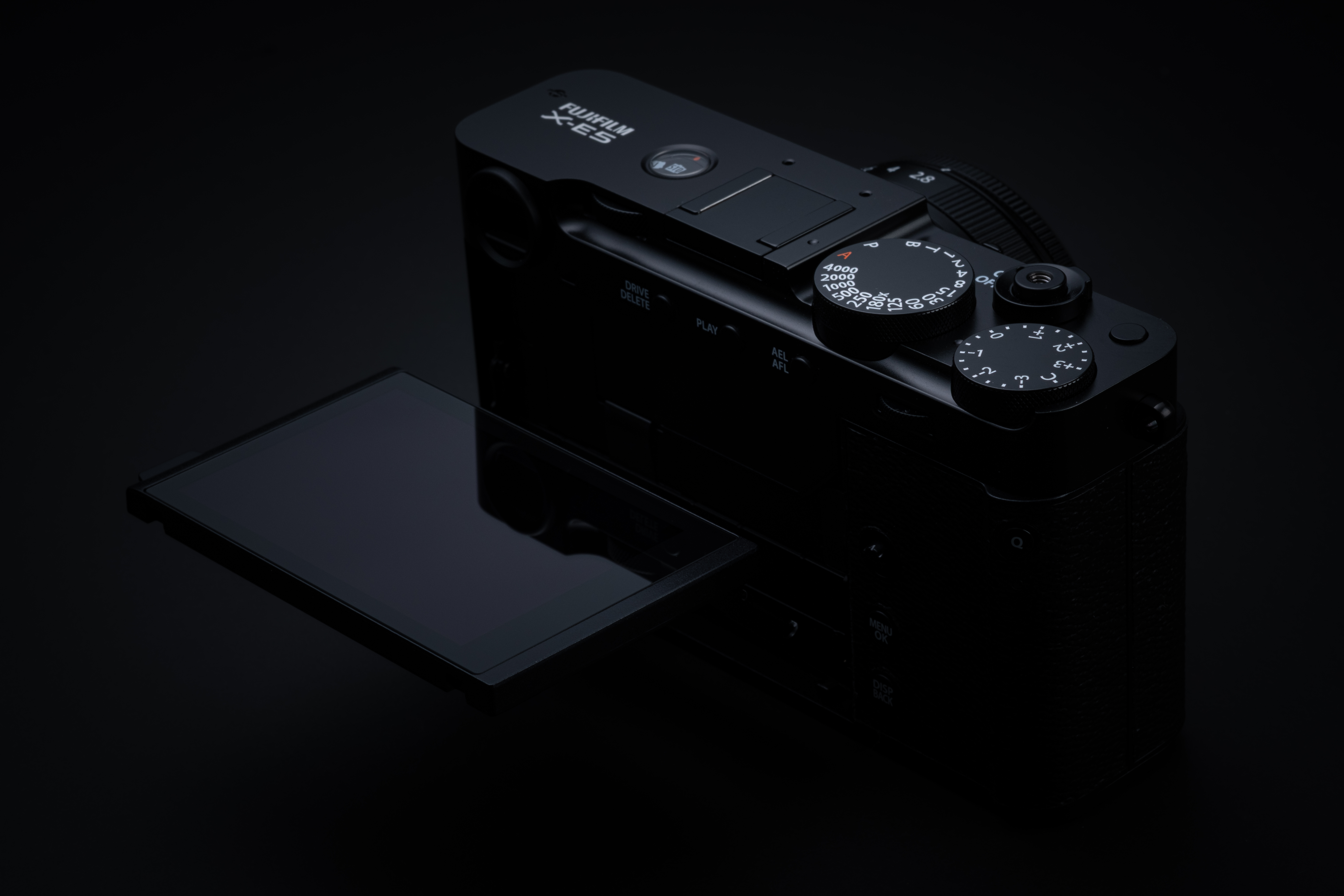 Schwarze Fujifilm X-E5 spiegellose Systemkamera aus einer schrägen Rückansicht vor tiefschwarzem Hintergrund, mit nach links voll ausgeklapptem Touchscreen-Display sowie Sicht auf die analogen Einstellräder und den gravierten weißen Fujifilm X-E5 Schriftz
