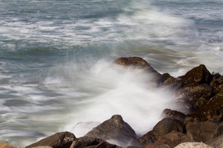 Langzeitbelichtung von Meereswellen, die mit dynamischer Bewegungsunschärfe und weißer Gischt an einer felsigen Küste brechen, zur Veranschaulichung von fließendem Wasser in der Landschaftsfotografie.