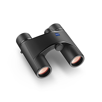 Schwarzes Zeiss Victory Pocket Fernglas aus einer schrägen Perspektive von oben, flach liegend mit sichtbaren rötlich vergüteten Frontlinsen und dem blauen Zeiss-Logo auf der Gehäuseoberseite, freigestellt vor weißem Hintergrund mit weichem Schatten.