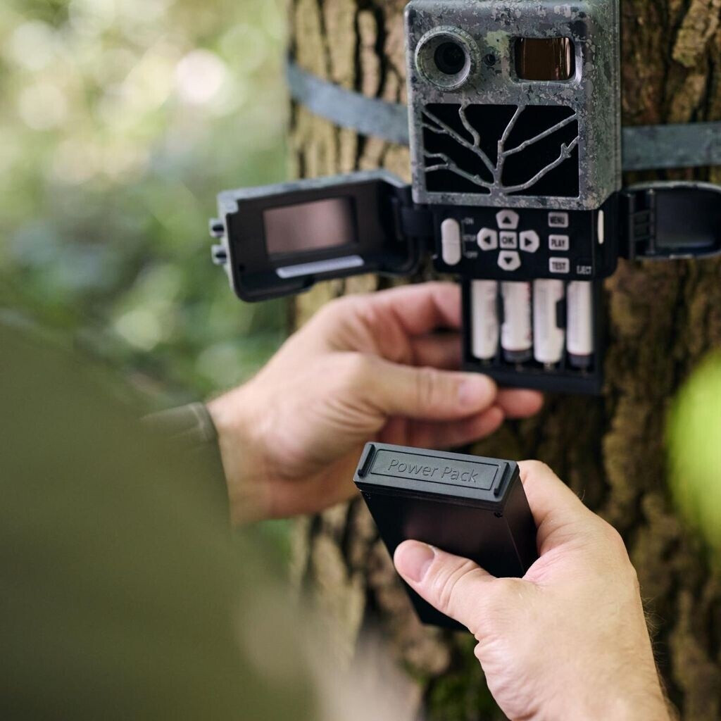 Eine Nahaufnahme zeigt die Hände einer Person im Freien, die ein schwarzes Batteriemagazin mit der Aufschrift „Power Pack“ in das geöffnete Batteriefach einer an einem Baumstamm befestigten ZEISS Secacam Wildkamera im Tarnmuster einsetzen. Das Tastenfeld
