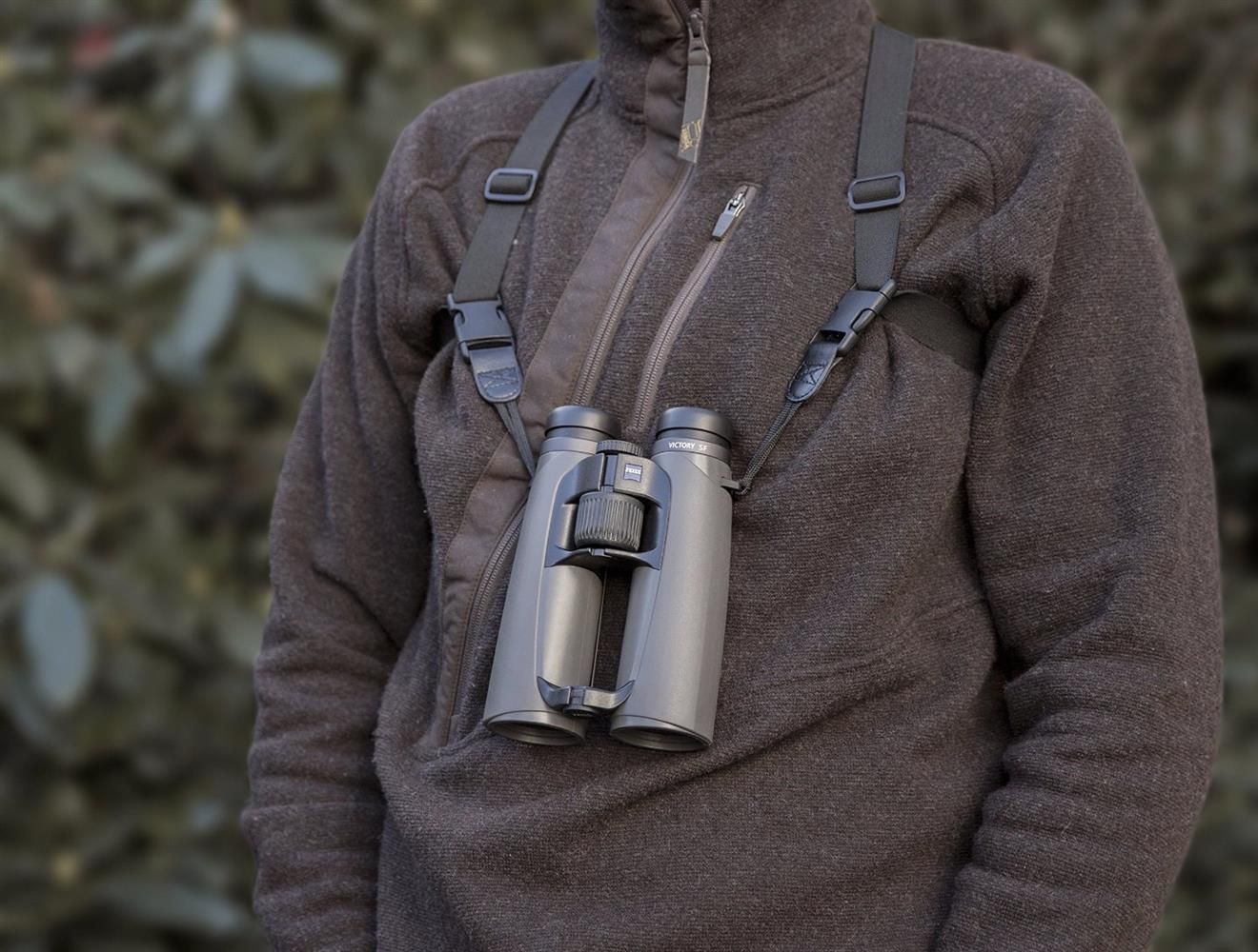Person mit brauner Fleecejacke trägt ein Fernglas an einem schwarzen Tragegurt vor der Brust; im Hintergrund ist unscharfe Vegetation zu sehen.