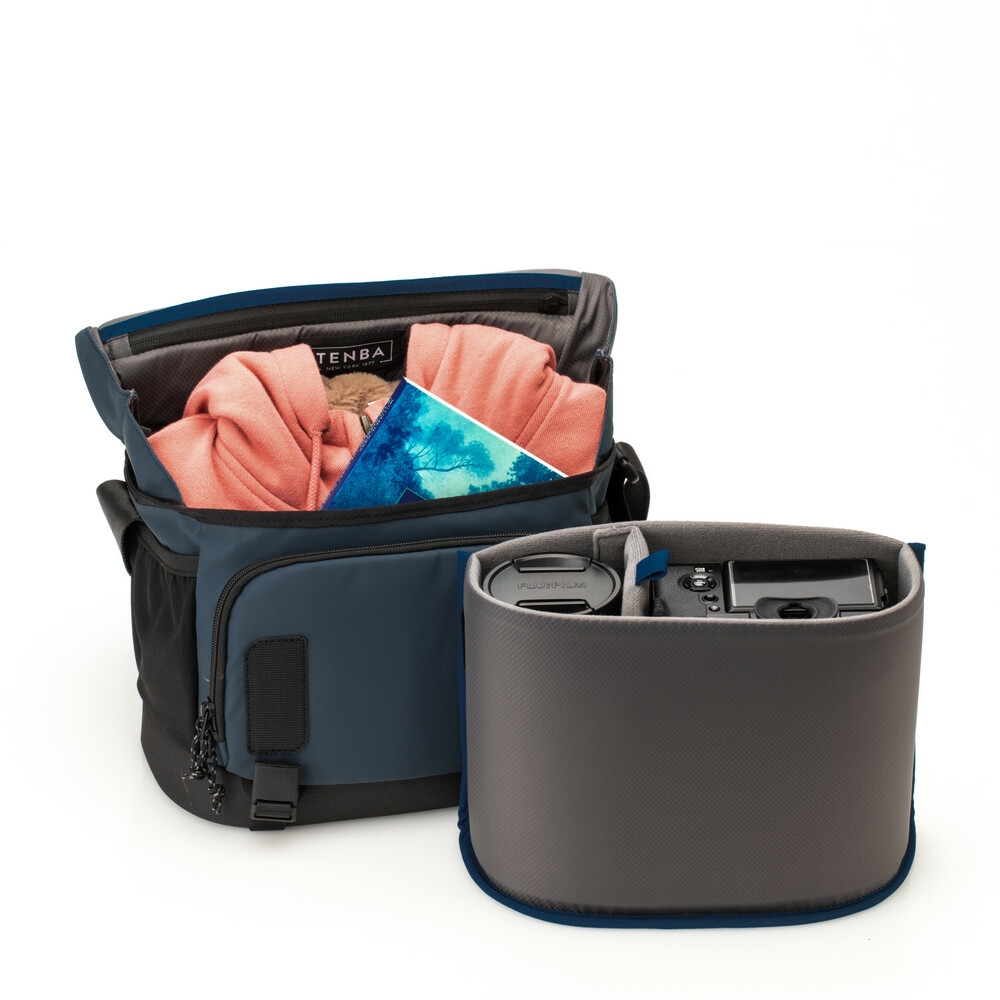 Geöffnete Tenba DNA Messenger Bag in Blau und Schwarz vor weißem Hintergrund mit herausgenommenem, grauem Kameraeinsatz, der eine Kamera und ein Objektiv enthält, während die Tasche als Freizeittasche mit Kleidung genutzt wird.