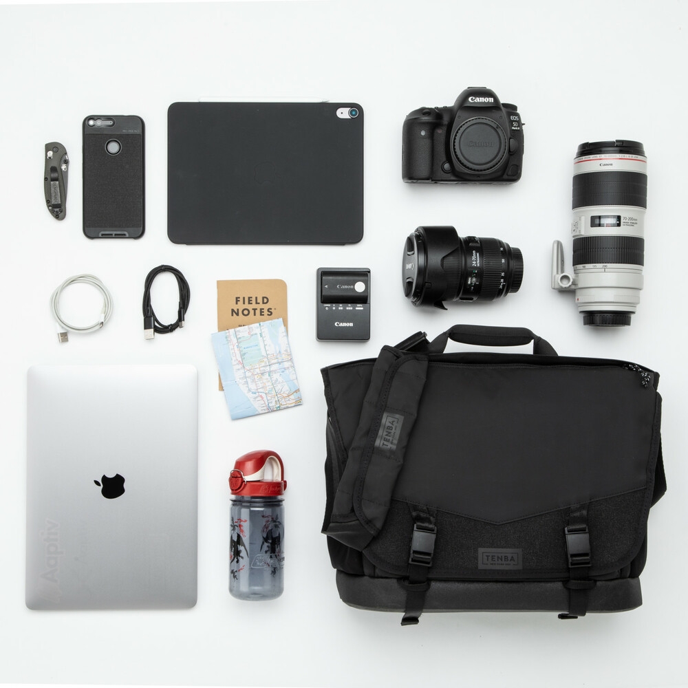 Draufsicht (Knolling) einer schwarzen Tenba DNA Messenger Bag Kameratasche vor weißem Hintergrund mit ausgebreitetem Equipment wie einer Canon DSLR-Kamera, zwei Objektiven, einem Apple MacBook, Notebook und Zubehör.