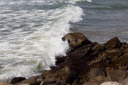 Naturaufnahme einer sich brechenden Meereswelle, die mit kurzer Belichtungszeit fotografiert wurde, sodass Wassertropfen und weiße Gischt an dunklen Küstenfelsen scharf eingefroren wirken.