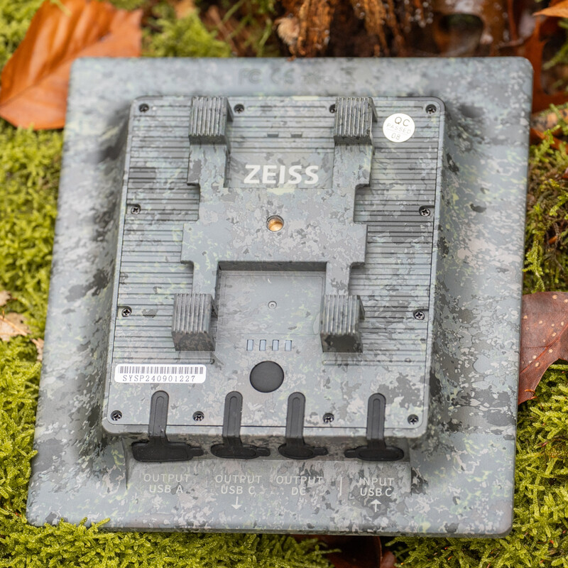 Die Rückseite des quadratischen ZEISS Secacam Solar Panels, flach auf moosigem Waldboden mit herbstlichen Blättern fotografiert. Das wetterfeste Gehäuse im grau-grünen Camouflage- beziehungsweise Rinden-Design zeigt in der Mitte eine erhabene H-förmige Mo