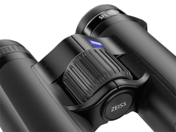 Eine Nahaufnahme des Fokussierrads eines schwarzen ZEISS Victory SFL Fernglases vor weißem Hintergrund. Im Mittelpunkt steht das große, vertikal geriffelte Fokussierrad der Brücke. Die vordere Abdeckung der Mittelachse trägt die weiße Aufschrift „ZEISS“,