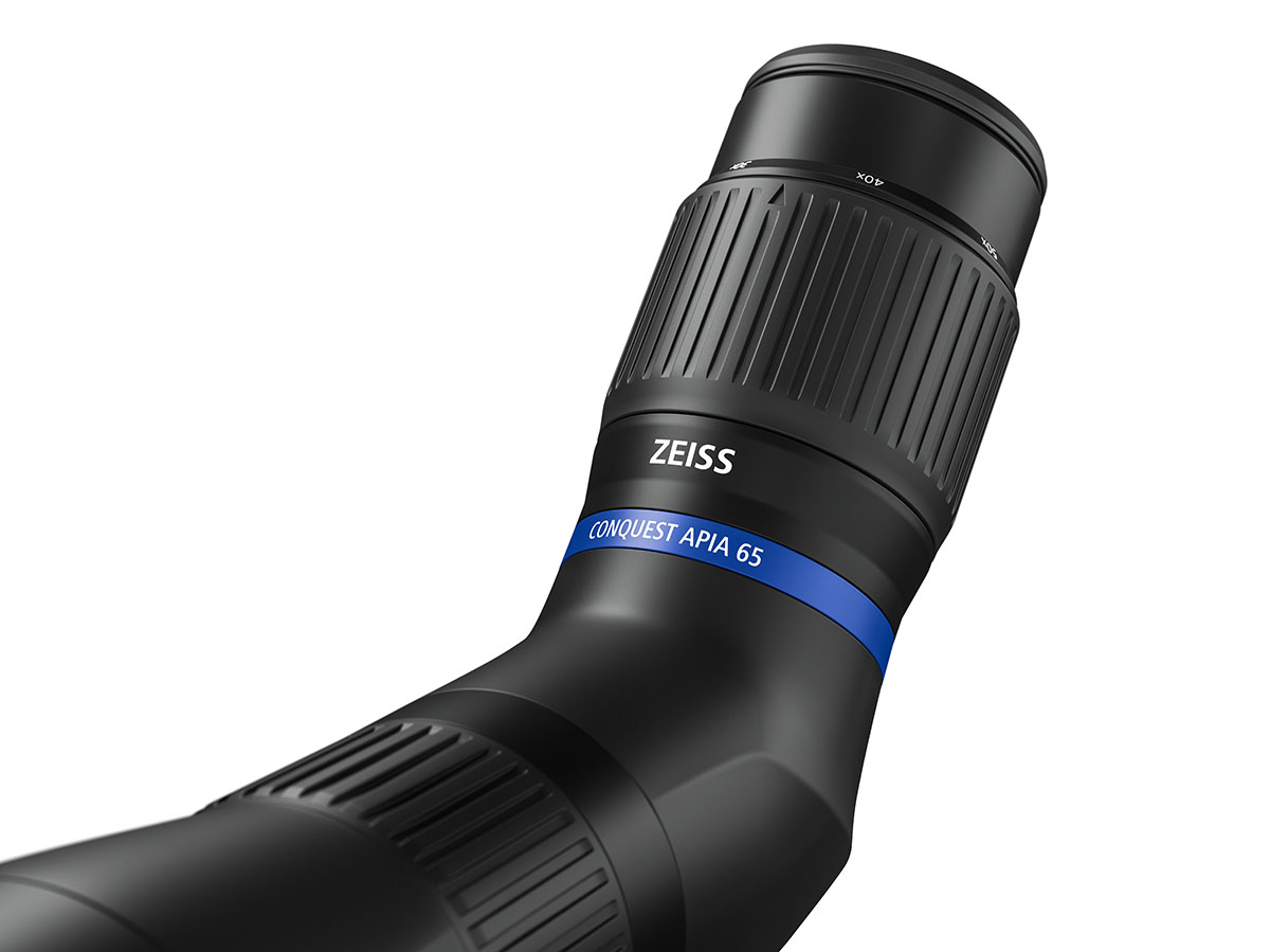 Eine Nahaufnahme des Okulartubus eines schwarzen ZEISS Conquest Apia 65 Spektivs vor weißem Hintergrund. Am Gehäuse ist die weiße Aufschrift „ZEISS“ zu sehen, direkt gefolgt von einem markanten blauen Ring mit dem weißen Schriftzug „CONQUEST APIA 65“. Der
