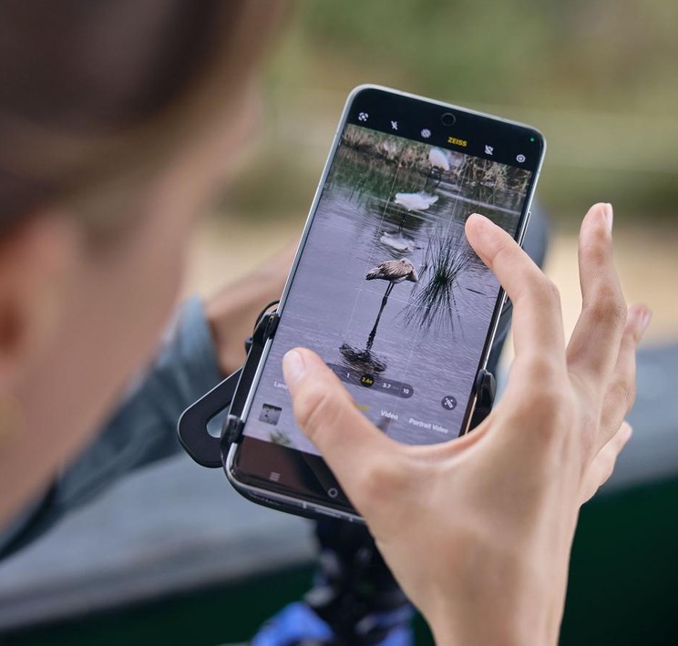 Eine Nahaufnahme über die Schulter einer Frau, die ein Smartphone bedient, das mithilfe des robusten ZEISS Digiscoping Phone Adapters an einem Spektiv oder Fernglas montiert ist [image_iMiezA.png]. Auf dem hochauflösenden Handydisplay ist das vergrößerte