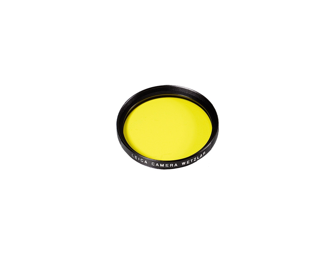 Perspektivische Draufsicht eines runden gelben Leica Kamera-Objektivfilters mit schwarzem Metallrahmen und weißer Randbeschriftung auf weißem Hintergrund.