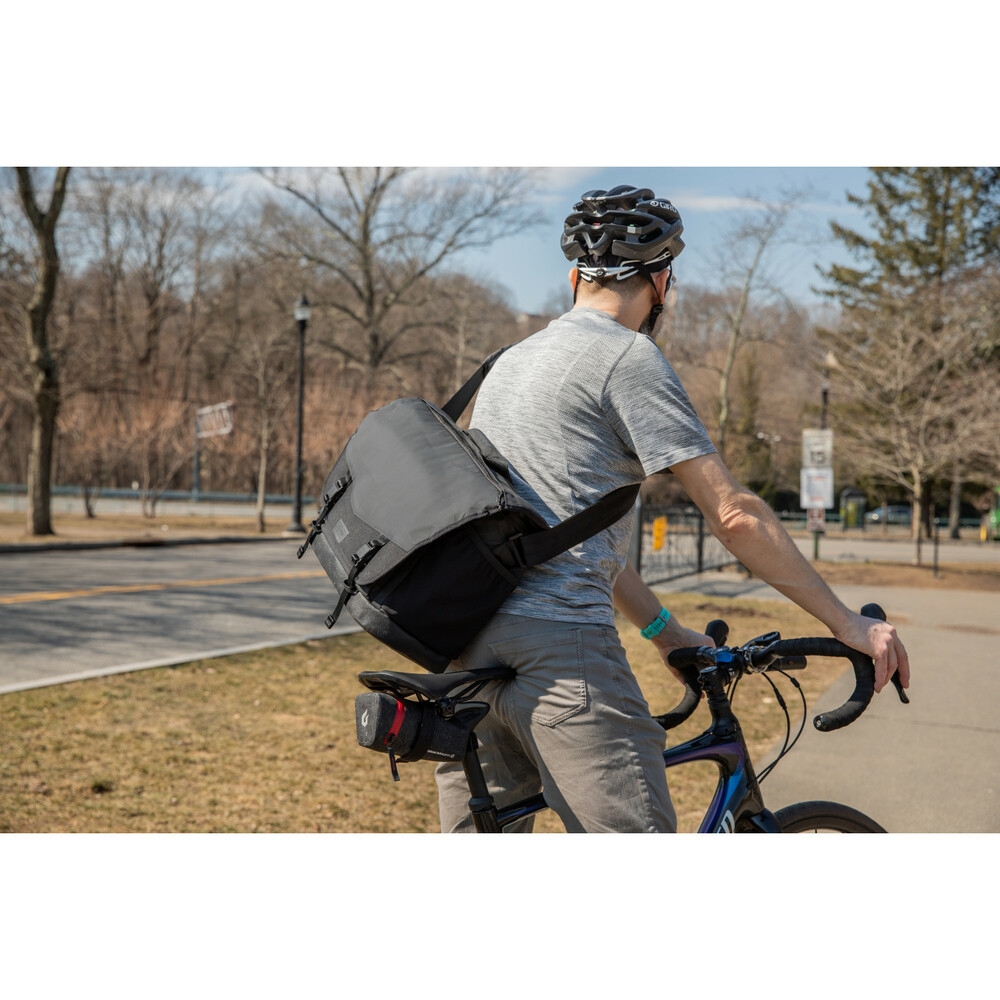 Ein Radfahrer von hinten mit Helm, der eine graue Tenba DNA Messenger-Kameratasche auf dem Rücken trägt und auf einem Rennrad im Freien sitzt, mit Bäumen im Hintergrund.