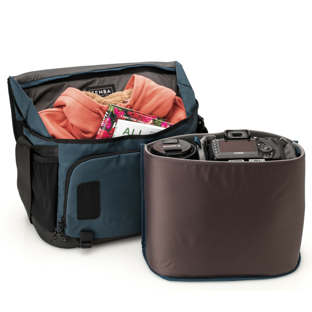 Geöffnete blau-schwarze Tenba DNA Messenger Bag Kameratasche vor weißem Hintergrund mit herausgenommenem, grauen Kameraeinsatz, der eine DSLR-Kamera und ein Objektiv schützt, während das Hauptfach der Tasche mit einem Pullover und Magazin befüllt ist.