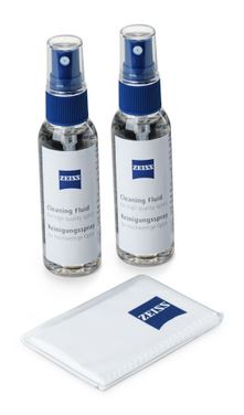 Ein dreiteiliges ZEISS Optik-Reinigungsset bestehend aus zwei Sprühflaschen und einem Reinigungstuch, leicht schräg von oben auf einem weißen Hintergrund fotografiert. Im oberen Bereich stehen nebeneinander zwei transparente 60-ml-Flaschen mit flüssigem Z